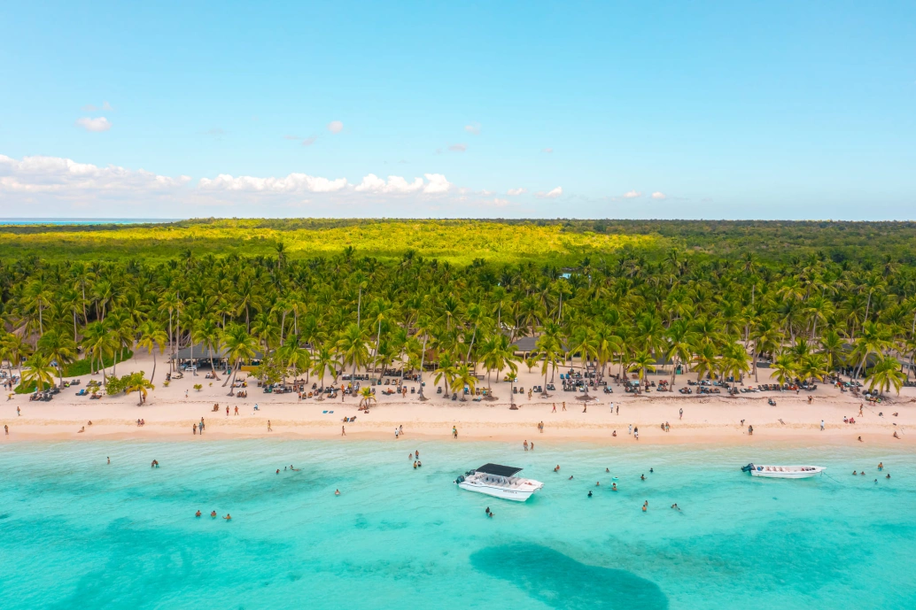 Vista aérea de uma praia em Punta Cana. Há diversos turistas na areia e no mar de cor azul turquesa. Ao fundo, coqueiros e vegetação densa