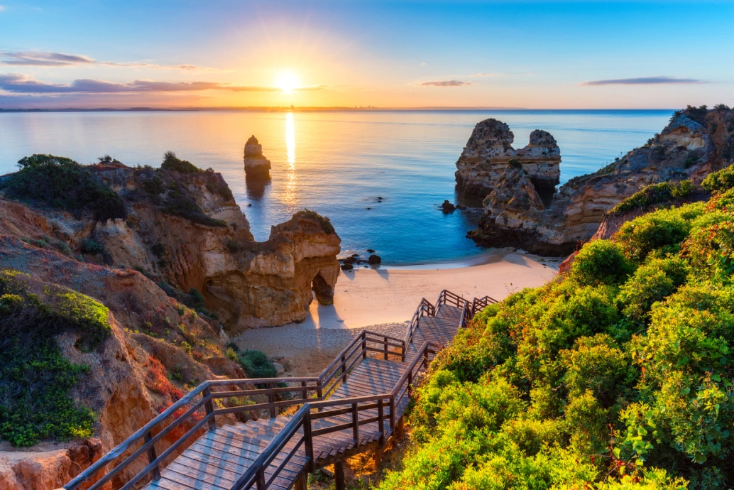 Praia do Camilo no Algarve, Portugal. Mar azul turquesa contornado por falésias e vegetação. Belíssimo pôr do sol ao fundo. Uma escadaria de madeira leva até a areia da praia.