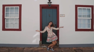 Garota posando de braços abertos à frente de casa colonial com porta azul escuro em rua histórica
