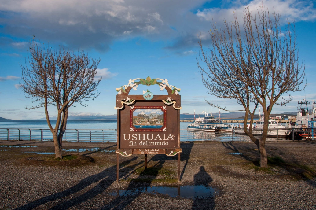 Placa Ushuaia, Argentina, com os dizeres “fim del mundo”. Ao fundo, a paisagem típica argentina, barcos, um vasto espelho d'água e as montanhas encerrando o horizonte