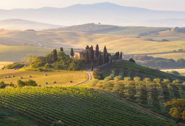 Bela vista da Toscana, na Itália, durante a primavera, com campos de prados e colinas ao pôr do sol. Bem ao centro da imagem, há uma casa típica da região, adornada por ciprestes.