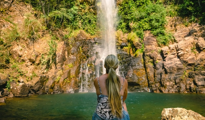Mulher sentada de costas em frente à Cachoeira Serra Azul em dia ensolarado