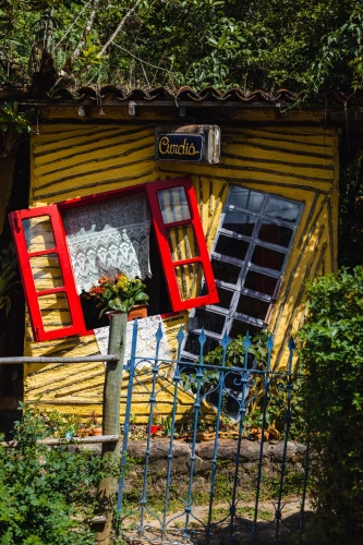 Pequena e curiosa casinha com fachada amarela, com janela e porta inclinadas para os lados. Há uma grade azul na frente e vegetação ao fundo, em Lavras Novas