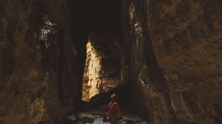 Grandes paredões de cânion levam a uma abertura na formação rochosa expondo uma fresta de luz do dia. Ao centro da imagem, um homem olha para cima.
