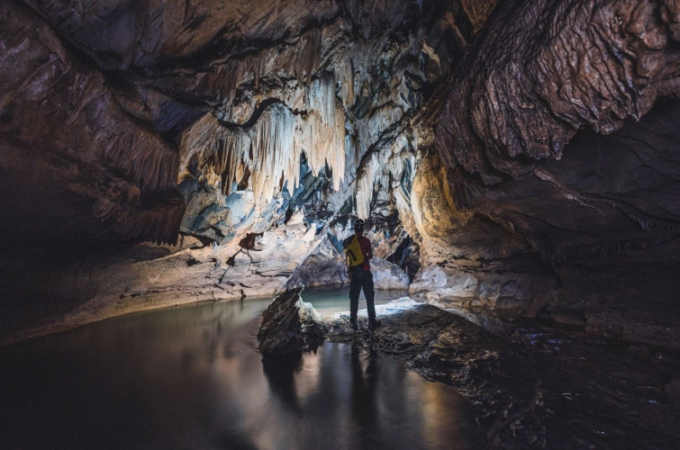 Homem de capacete com lanterna em pé em meio às estruturas de uma caverna escura, iluminando as estalactites
