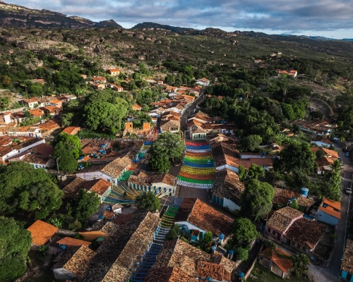 Vista aérea de um povoado com várias casas e seus telhados e diversas árvores pelas ruas, além de uma montanha cercando a região.