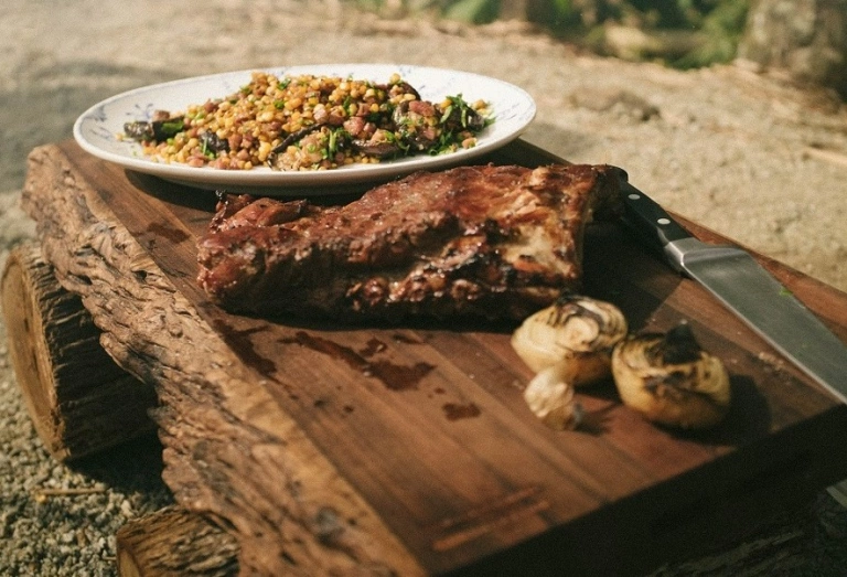 Uma tábua de madeira comporta uma costela suína assada ao lado de um prato de vegetais para acompanhamento