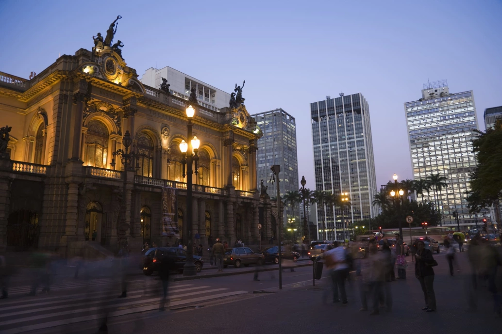 Construção em estilo clássico com luzes acesas na fachada. Há movimento de pessoas e carros em frente ao local, e prédios modernos ao fundo na cidade de São Paulo