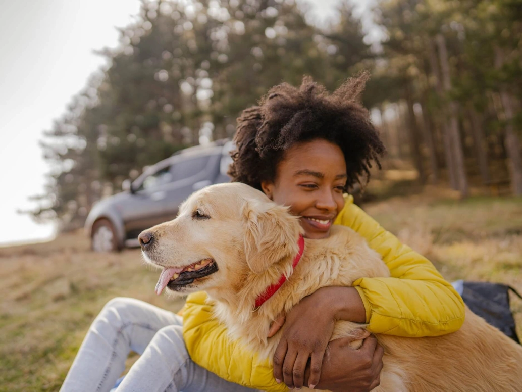 Mulher negra usando calça jeans e uma blusa de frio amarela abraça cachorro. Ambos estão sentados na grama com um carro e árvores ao fundo.