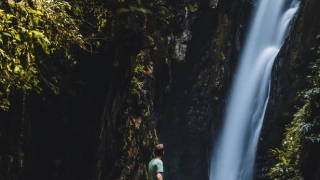 Homem em pé em cima de uma pedra observando uma cachoeira em meio a uma vegetação densa