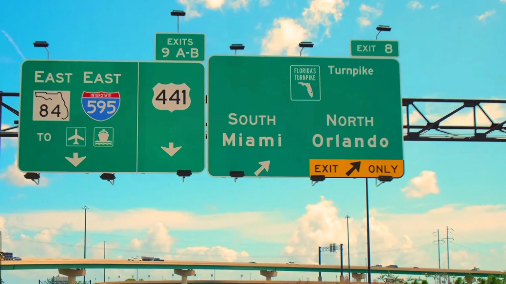 Foto de placas verdes de sinalização de trânsito indicando a rota Miami - Orlando, saídas, localização da rodovia, aeroporto e guarda costeira. Ao fundo, céu azul com algumas nuvens brancas.