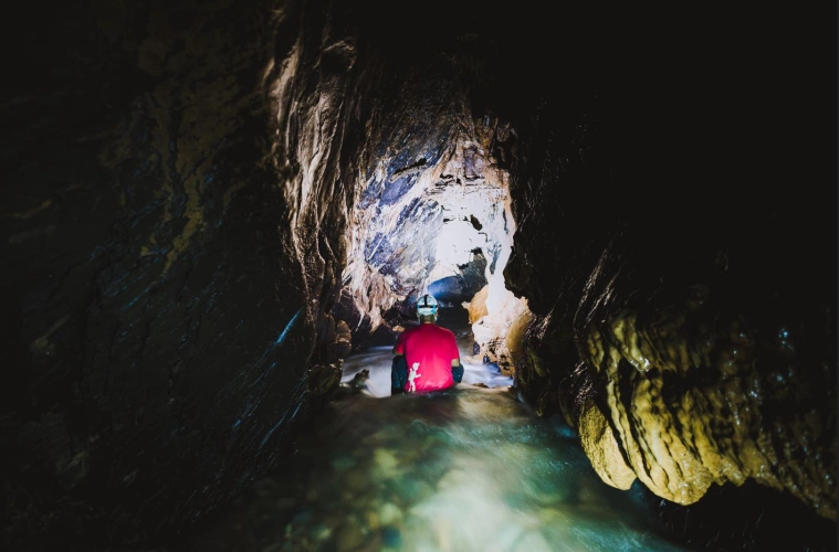 Homem de capacete com lanterna sentado próximo a um poço às estruturas de uma caverna escura