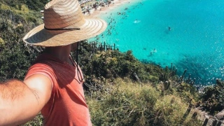 Homem tira foto de si mesmo em frente à praia de água cristalina em dia ensolarado