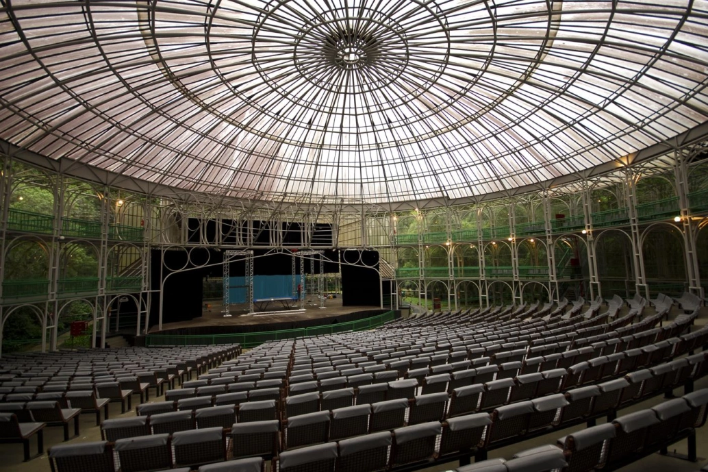 Vista interna de um espaço para espetáculos feito em estrutura metálica. A foto mostra os assentos da plateia, o palco e o teto do local