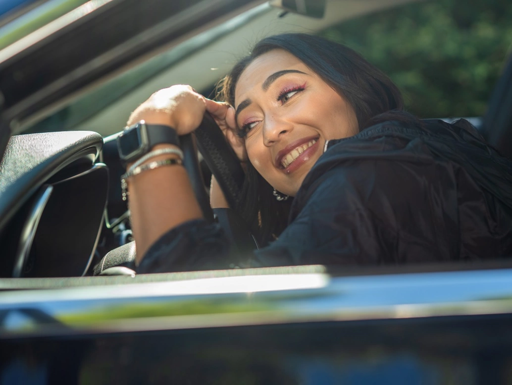 Uma mulher de cabelos pretos  sorrindo e abraçando um volante de carro