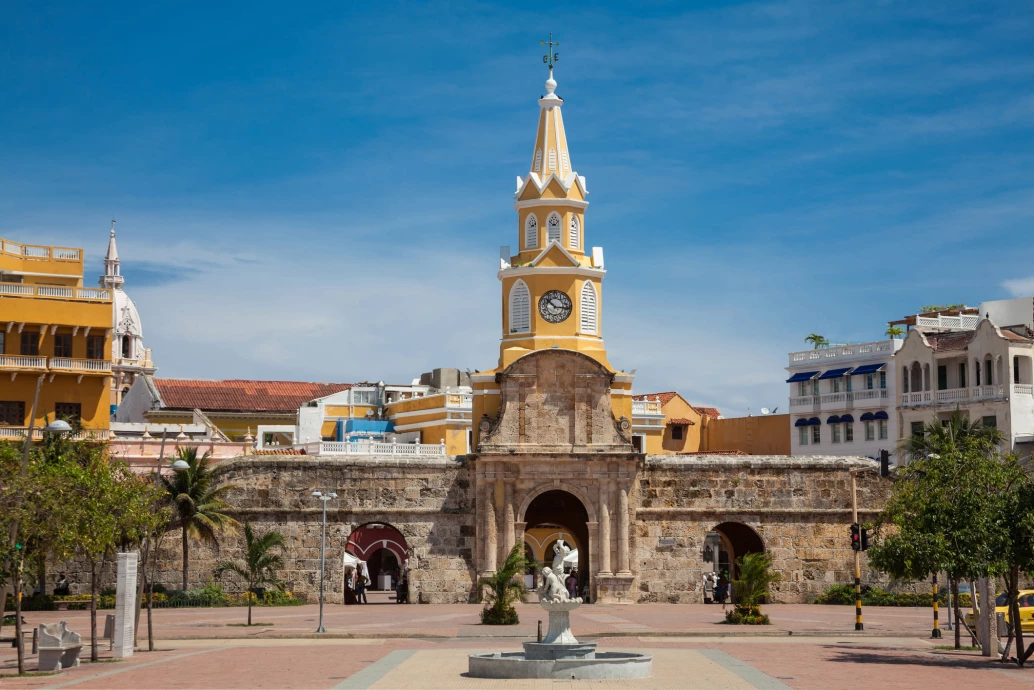 Vista frontal de uma torre com relógio no topo, localizada atrás de uma muralha em dia ensolarado na cidade de Cartagena, na Colômbia.