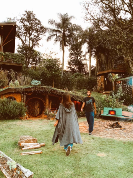 Um homem e uma mulher caminham num pátio onde há uma estrutura de madeira e árvores. O fundo apresenta uma casa subterrânea semelhante à do filme "O Hobbit" na cidade de Jundiaí, SP.