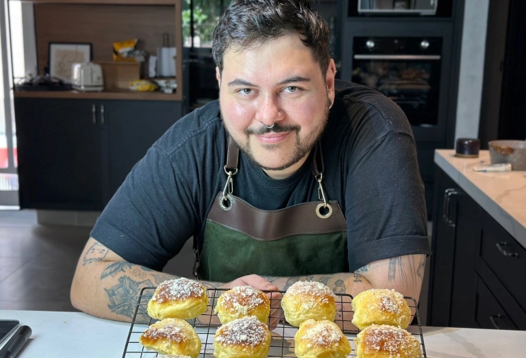 Homem em uma cozinha apoiado em mesa contendo pães amarelos