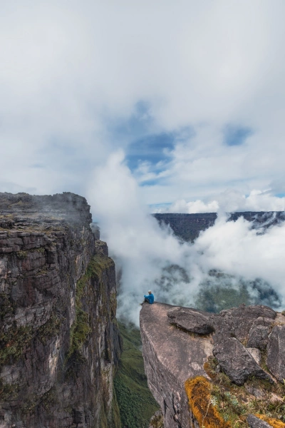 Homem sentado na beirada de uma pedra a mais de 2 mil metros de altitude. Há neblina e paredões rochosos ao fundo