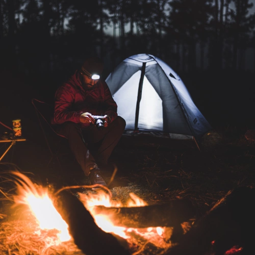 Homem em frente a uma fogueira em meio à natureza. Barraca de acampamento ao fundo