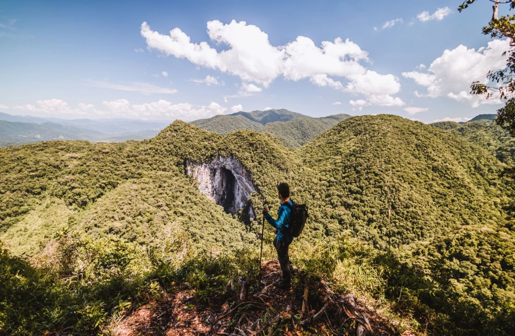 Homem contempla vasta cadeia de montanhas coberta por vegetação, em belo dia claro. Ao centro, se destaca uma abertura na paisagem que desvenda uma gruta, destino do explorador.