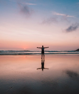 Homem de braços abertos em direção ao pôr do sol numa praia deserta em fim de tarde.