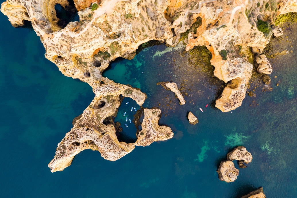Vista aérea da Ponta da Piedade no Algarve, Portugal. As formações rochosas se estendem mar adentro, com coloração dourada que contrasta com o azul-esverdeado da água.