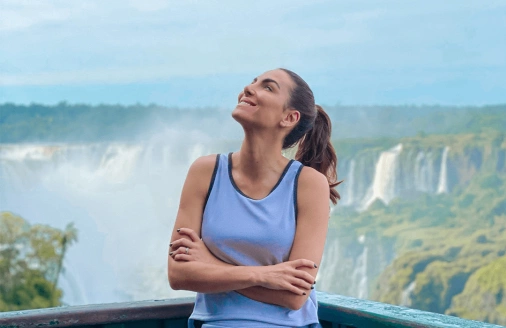 Mulher de braços cruzados e encostada em uma grade sorri e olha emocionada para o céu. Ao fundo, as quedas d’água das Cataratas do Iguaçu.