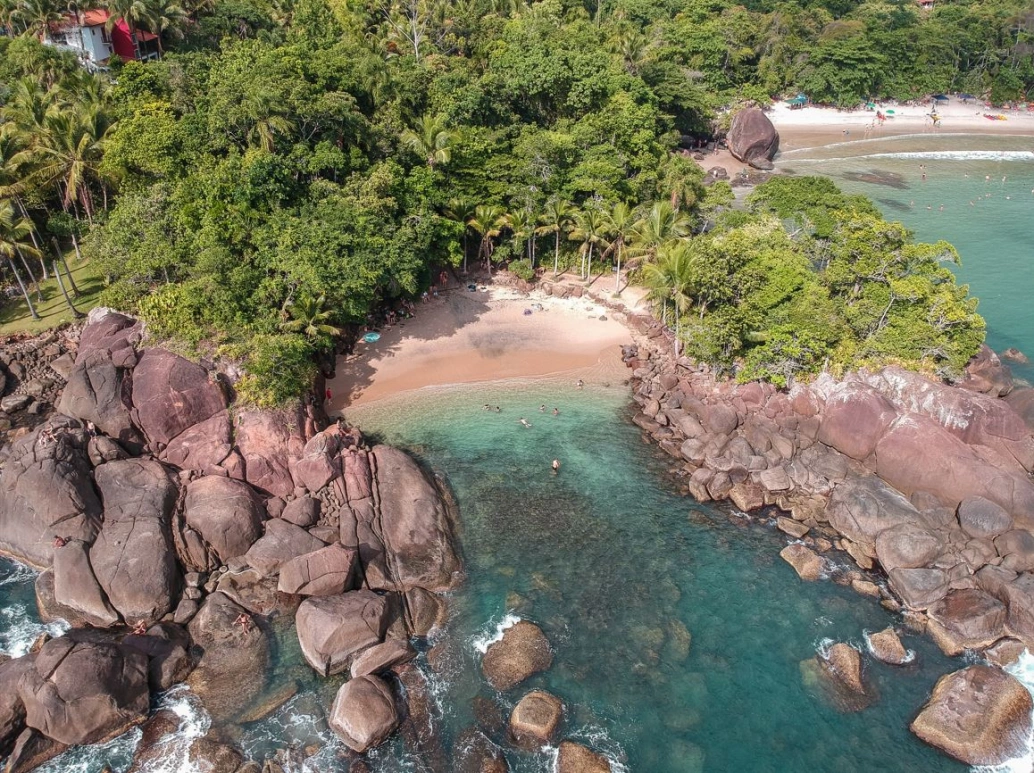 Vista aérea de praia com mar verde-esmeralda, cercada por grandes pedras e vegetação selvagem. Há alguns banhistas na água e outros na sombra dos coqueiros