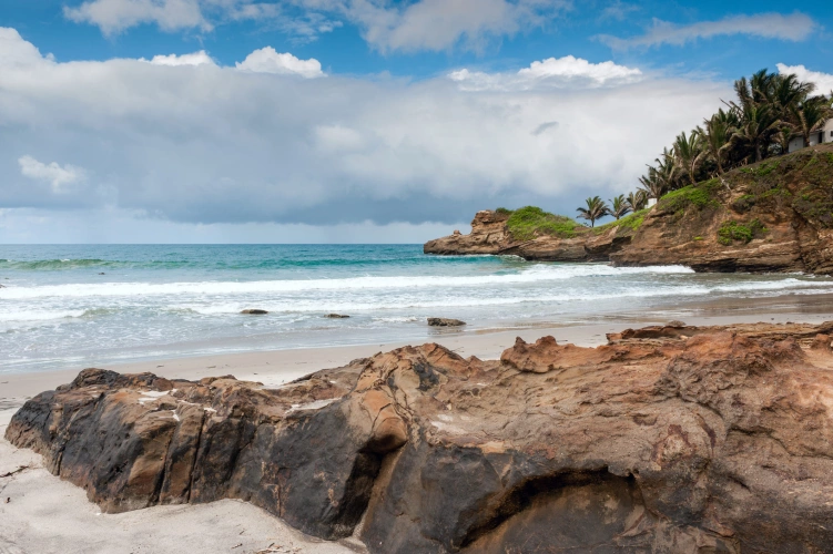 Praia do litoral equatoriano. No plano frontal, uma rocha em meio à areia. Ao fundo, mar de águas cristalinas e céu azul com nuvens