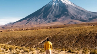 Homem se destaca olhado para vulcão em meio à vegetação desértica.