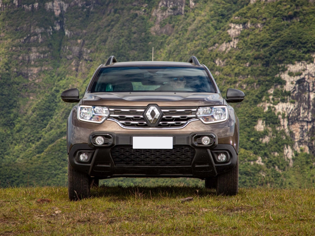 Frente de um Renault Duster marrom estacionado em um campo com montanhas ao fundo.