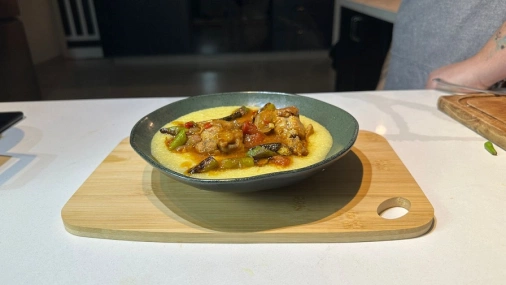 um prato de cerâmica preto servido com polenta cremosa, coberta com pedaço da cozinha do frango, imersas em um molho dourado, junto com pedaços de quiabo. O prato está sob uma tábua de madeira clara.