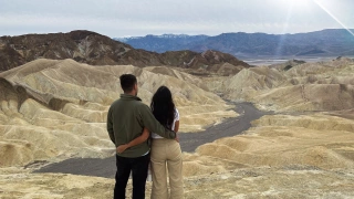 Casal abraçado observando montanhas rochosas com variados tons de amarelo e marrom. Ao fundo, mais montanhas e raios de sol em meio ao céu nublado