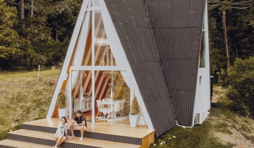 Casal sentado à frente de casa em formato triangular e com janelas de vidro em região arborizada