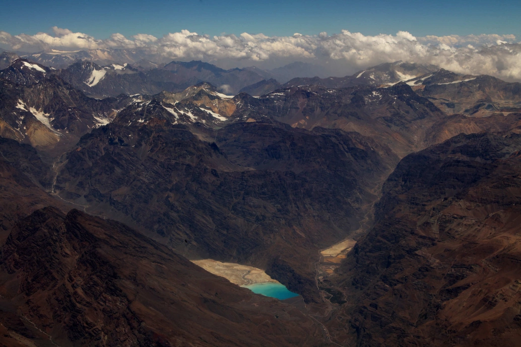 Extensas formações rochosas marrons formam a Cordilheira dos Andes. Ao fundo, a neve cobre os últimos picos à vista, num dia carregado de nuvens.