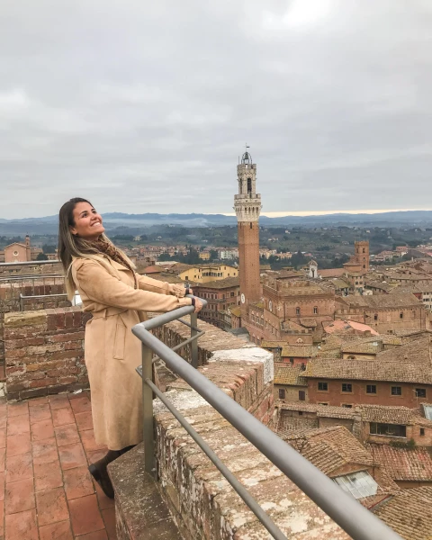 Jovem mulher brasileira, posa para foto em parapeito que dá uma bela vista para Siena, Toscana. Ao fundo, vemos as construções típicas medievais locais.
