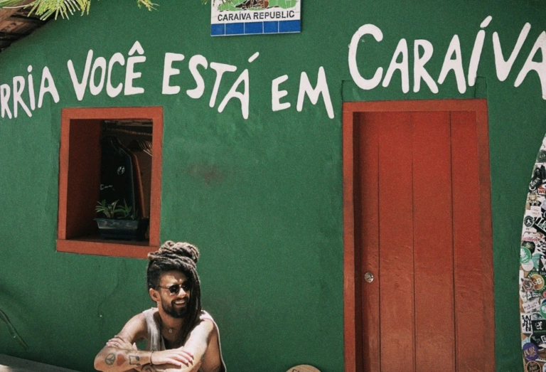 Músico Gabriel Elias sentado em frente à casa de fachada na cor verde com os dizeres "sorria você está em caraíva"