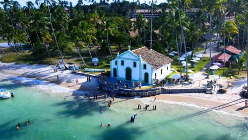 Vista aérea da Praia dos Carneiros. Há uma pequena capela à beira mar, onde estão alguns turistas. Ao fundo, muitos coqueiros e alguns guarda-sóis de praia