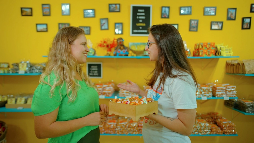 Duas mulheres conversam sorrindo em uma loja de produtos comestíveis, a bala Bananinha, com prateleiras e quadros ao fundo. Uma delas segura um recipiente com as balas.