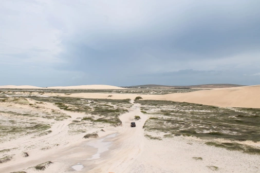 Vista aérea de carro passando por estrada em meio às dunas de areia com leve vegetação rasteira