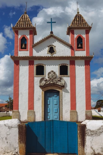 Fachada de igreja com parede branca, colunas vermelhas e porta azul em Lavras Novas. Há uma porteira azul no plano frontal, uma cruz no topo da igreja e céu azul com nuvens ao fundo