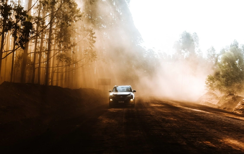 Carro em estrada de terra, à frente da poeira levantada por seu movimento. Feixes de luz passam por entre as árvores da encosta da estrada.