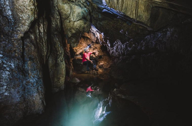 Homem de capacete com lanterna sentado em pedra, em meio às estruturas de uma caverna escura