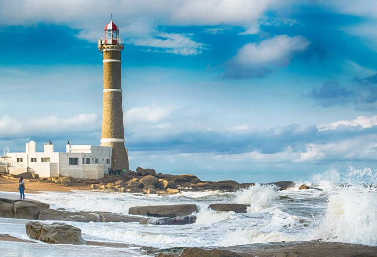 Praia no Uruguai, com água do mar batendo em algumas pedras. Ao fundo, um farol e céu azul com nuvens