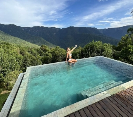 Mulher sentada na beira da piscina com braço estendido para o céu em dia ensolarado. Ao fundo, paisagem de cadeira de montanhas da Serra Catarinense.