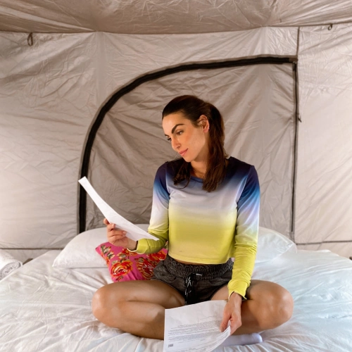 Mulher sentada em colchão dentro da barraca segurando e lendo alguns papéis