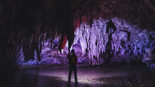 Homem em pé com luz ao redor, em meio às estruturas de uma caverna. Destaque para as estalactites no teto