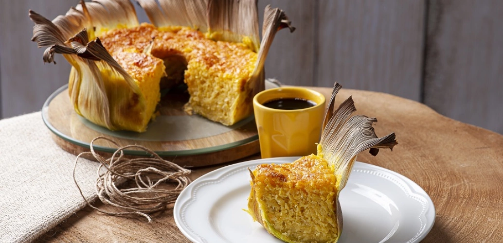 Uma fatia de bolo de milho sobre um pires ao lado de uma xícara de café e do restante do bolo sob uma mesa decorada