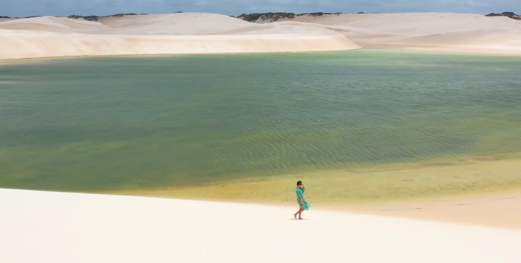 Mulher caminha por dunas de areia branca e enorme lagoa de águas esverdeadas